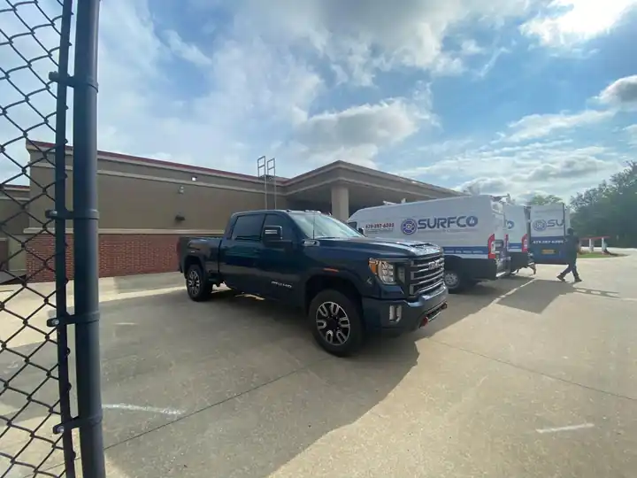Three white Surfco vans and a black pickup truck parked in front of a brick commercial building under a wide sky