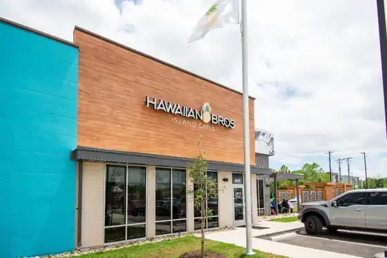 Hawaiian Bros Island Grill storefront with a teal wall, wood cladding, tall windows and a flagpole