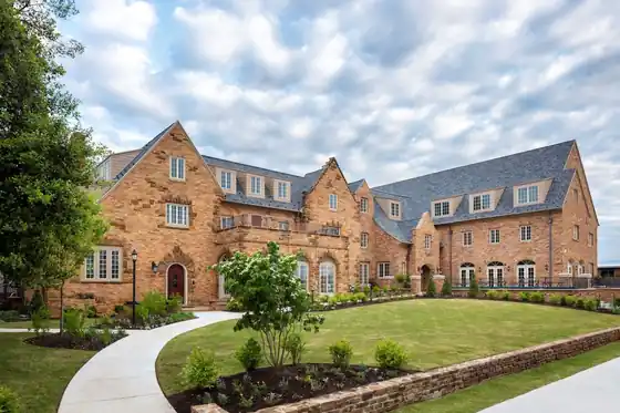 Stone and brick collegiate-style building with a slate roof, a curved walkway and a landscaped lawn under a cloudy sky