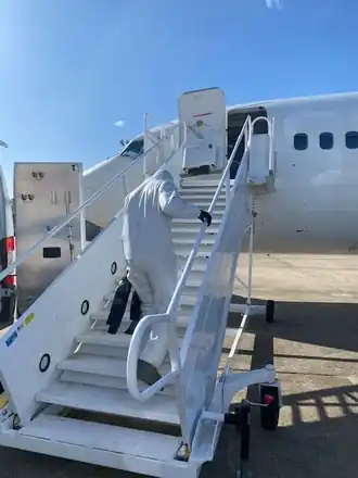 A technician in a white Tyvek suit climbing an aircraft's boarding stairs carrying a sprayer