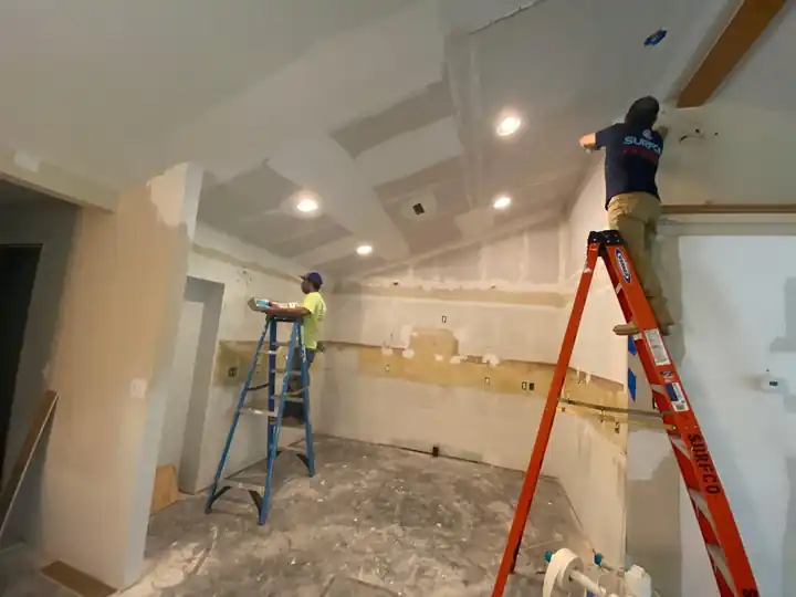 Two workers on orange ladders finishing drywall in a vaulted room