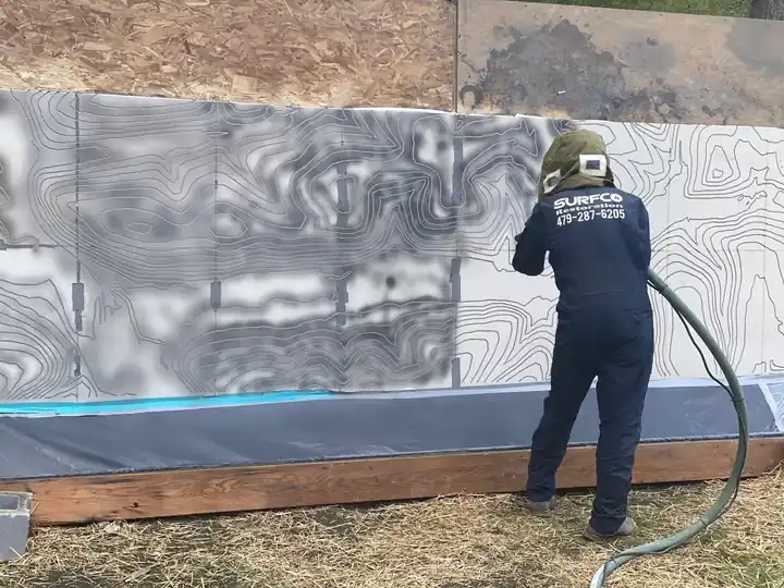 Surfco technician in navy coveralls blasting paint off a long plywood wall, seen from behind with the hose in hand