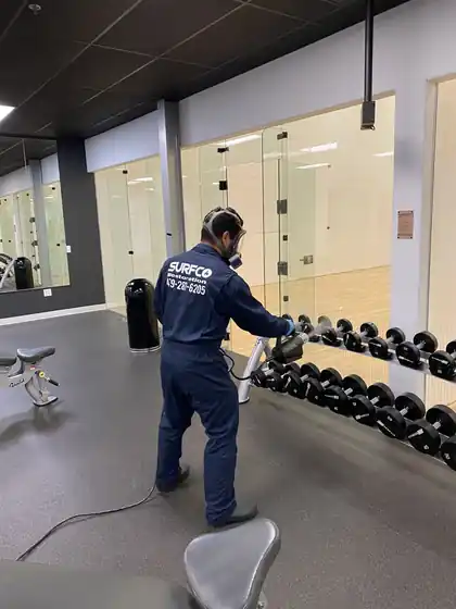 A Surfco technician in navy coveralls disinfecting a dumbbell rack in a glass-walled gym